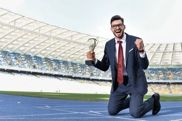 Heureux jeune homme d'affaires en costume debout sur les genoux sur la piste de course avec trophée au stade — Photo de stock