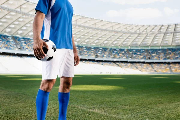 Vista recortada de jugador de fútbol profesional en uniforme azul y blanco con pelota en el campo de fútbol en el estadio - foto de stock