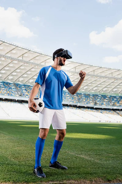 Professional soccer player in vr headset and blue and white uniform with ball yelling and showing yes gesture on football pitch at stadium — Stock Photo