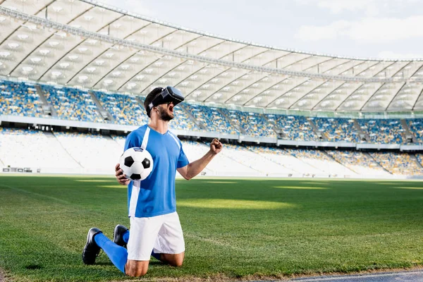 Jogador de futebol profissional em vr headset e uniforme azul e branco com bola de pé sobre os joelhos e mostrando sim gesto no campo de futebol no estádio — Fotografia de Stock