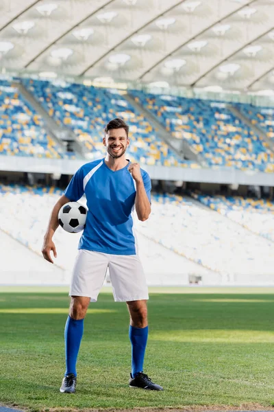 Jogador de futebol profissional feliz em uniforme azul e branco com bola mostrando sim gesto no campo de futebol no estádio — Fotografia de Stock