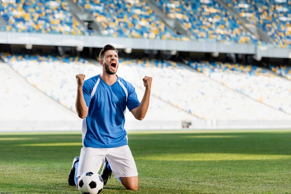 Jugador de fútbol profesional emocional en uniforme azul y blanco con pelota de pie sobre las rodillas en el campo de fútbol y mostrando sí gesto en el estadio - foto de stock