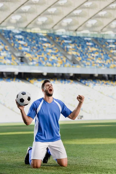 Emotionaler Fußballprofi in blau-weißer Uniform mit Ball, der auf Knien auf dem Fußballplatz steht und im Stadion Ja-Geste zeigt — Stockfoto