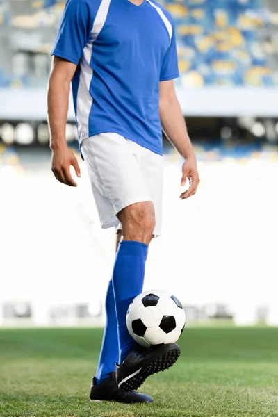 Vista recortada de jugador de fútbol profesional en uniforme azul y blanco con pelota en el campo de fútbol en el estadio - foto de stock