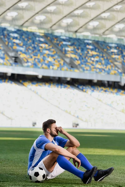 Triste footballeur professionnel en uniforme bleu et blanc assis avec ballon sur le terrain de football au stade — Photo de stock