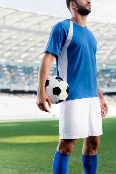 Vista recortada de jugador de fútbol profesional en uniforme azul y blanco con pelota en el campo de fútbol en el estadio - foto de stock