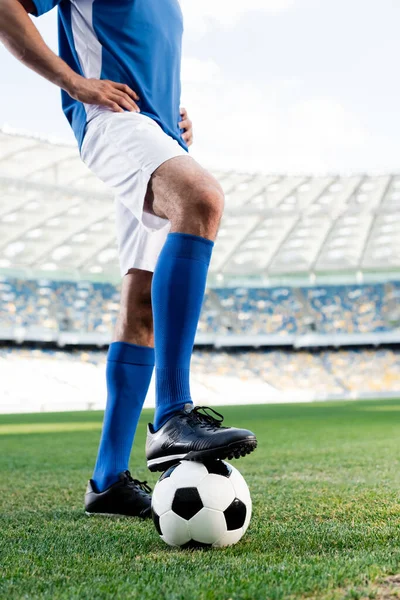 Vista cortada de jogador de futebol profissional em uniforme azul e branco com bola e mãos em quadris em campo de futebol no estádio — Fotografia de Stock