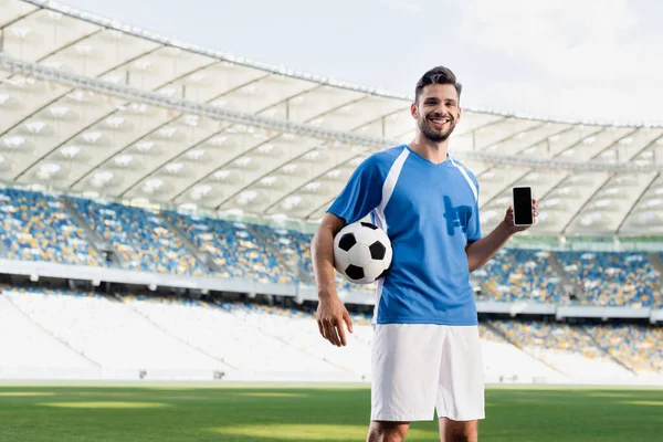 Smiling professional soccer player in blue and white uniform with ball showing smartphone with blank screen at stadium — Stock Photo