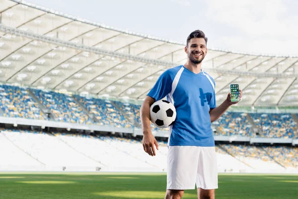 KYIV, UCRANIA - 20 de junio de 2019: jugador de fútbol profesional sonriente en uniforme azul y blanco con bola que muestra el teléfono inteligente con aplicaciones de iphone en el estadio - foto de stock