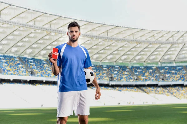 KYIV, UCRANIA - 20 de junio de 2019: futbolista profesional en uniforme azul y blanco con pelota mostrando smartphone con aplicación de youtube en el estadio - foto de stock