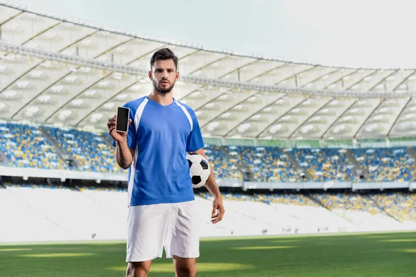 Professional soccer player in blue and white uniform with ball showing smartphone with blank screen at stadium — Stock Photo