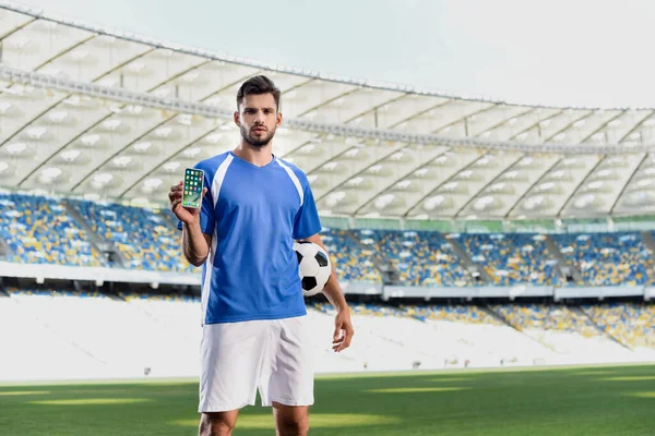 KYIV, UCRANIA - 20 de junio de 2019: futbolista profesional en uniforme azul y blanco con pelota que muestra el teléfono inteligente con aplicaciones de iphone en el estadio - foto de stock