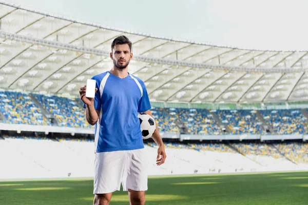 Jogador de futebol profissional em uniforme azul e branco com bola mostrando smartphone com tela em branco no estádio — Fotografia de Stock