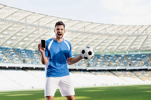 Calciatore professionista in divisa blu e bianca con palla che mostra smartphone con schermo bianco e grida allo stadio — Foto stock