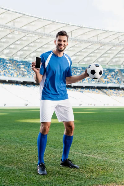 Jugador de fútbol profesional sonriente en uniforme azul y blanco con bola mostrando teléfono inteligente con pantalla en blanco en el estadio - foto de stock