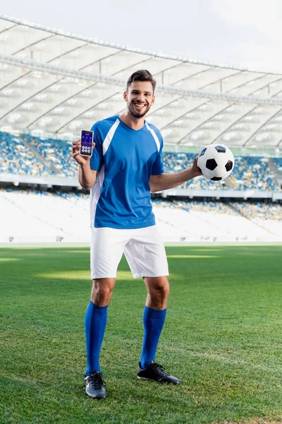Giocatore di calcio professionista sorridente in uniforme blu e bianca con palla che mostra smartphone con app sanitaria allo stadio — Foto stock