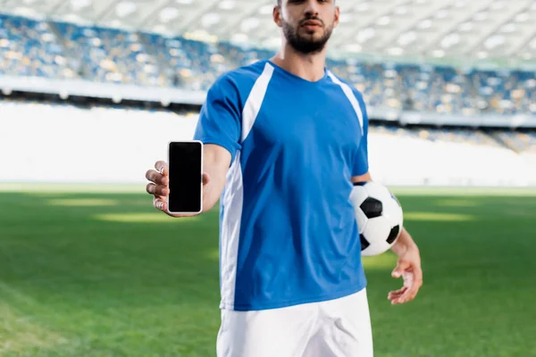 Cropped view of professional soccer player in blue and white uniform with ball showing smartphone with blank screen at stadium — Stock Photo