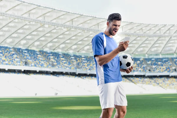 Emotional professional soccer player in blue and white uniform with ball using smartphone at stadium — Stock Photo