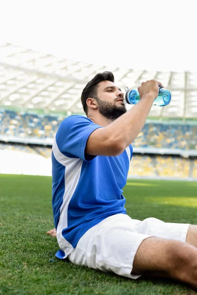 Calciatore professionista in divisa bianca e blu seduto sul campo da calcio e acqua potabile allo stadio — Foto stock