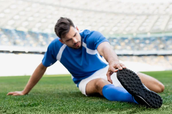 Selective focus of professional soccer player in blue and white uniform stretching on football pitch at stadium — Stock Photo