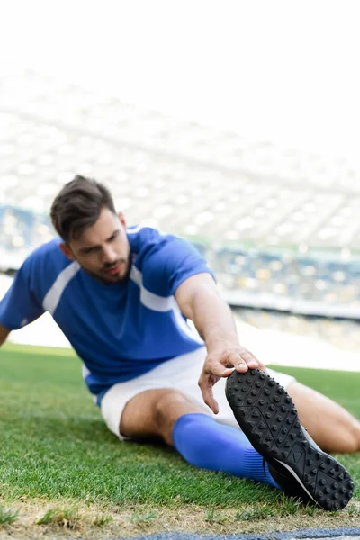 Enfoque selectivo del jugador de fútbol profesional en uniforme azul y blanco que se extiende en el campo de fútbol en el estadio - foto de stock