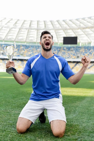 Professional soccer player in blue and white uniform with sports cup standing on knees on football pitch and shouting at stadium — Stock Photo