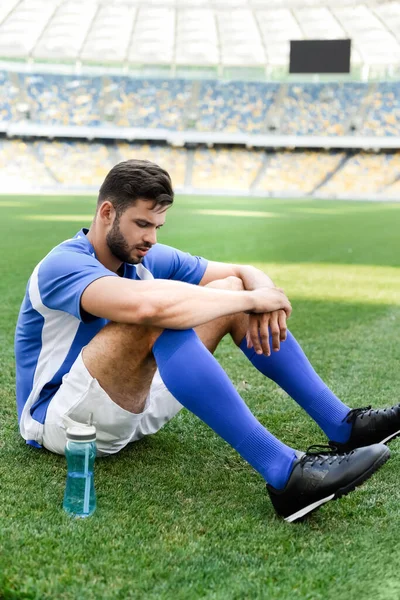 Professional soccer player in blue and white uniform sitting on football pitch with sports bottle at stadium — Stock Photo
