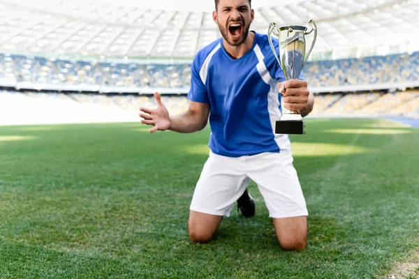 Профессиональный футболист в сине-белой форме со спортивным кубком, стоящим на коленях на футбольном поле и кричащим на стадионе — стоковое фото