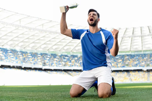 Futbolista profesional en uniforme azul y blanco con copa deportiva de pie sobre las rodillas en el campo de fútbol y gritando en el estadio - foto de stock
