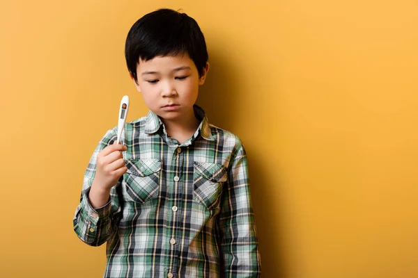 Trauriger asiatischer Junge hält Thermometer auf gelb — Stockfoto