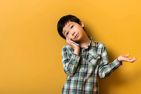 Un niño asiático escéptico que habla por teléfono inteligente en amarillo - foto de stock