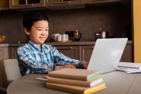 Chico asiático sonriente estudiando en línea con portátil y libros en casa durante la cuarentena. - foto de stock