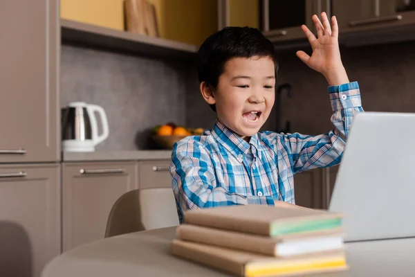 Эмоциональный мальчик с поднятой вверх рукой кричит и учится онлайн с ноутбуком дома во время самоизоляции — стоковое фото