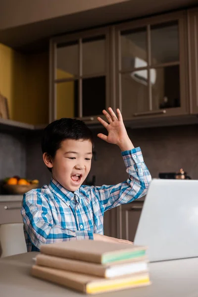 Jovencito asiático emocional con la mano levantando gritos y estudiando en línea con portátil en casa durante la cuarentena. - foto de stock