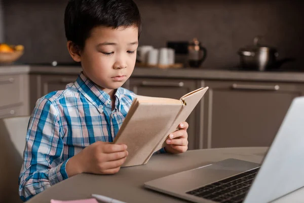Мальчик читает книгу и учится онлайн с ноутбуком дома во время самоизоляции — стоковое фото