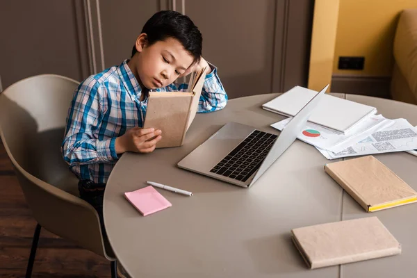 Asian child reading book and studying online with laptop at home during self isolation — Stock Photo