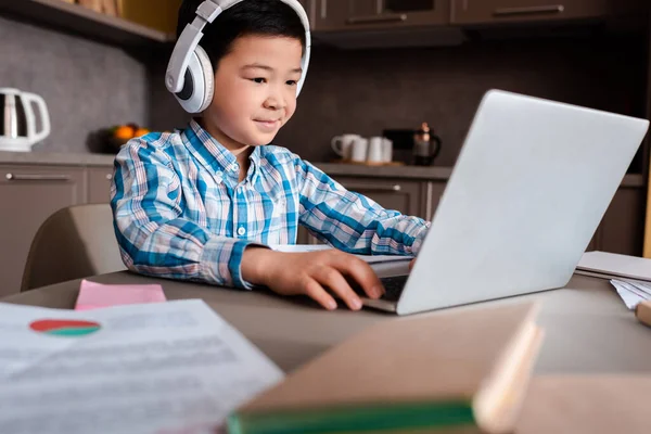 Chico asiático sonriente estudiando en línea con libros, portátiles y audífonos en casa durante la cuarentena. - foto de stock