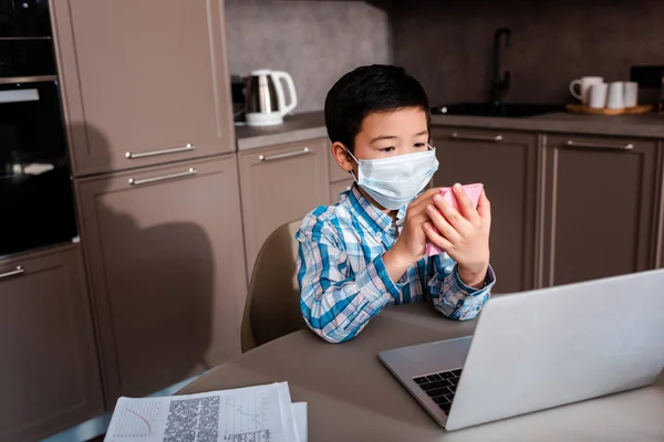 Adorable asian boy in medical mask writing and studying online with laptop at home during quarantine — Photo de stock