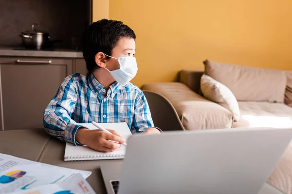 Ребенок в медицинской маске пишет и учится онлайн с ноутбуком дома во время карантина — стоковое фото