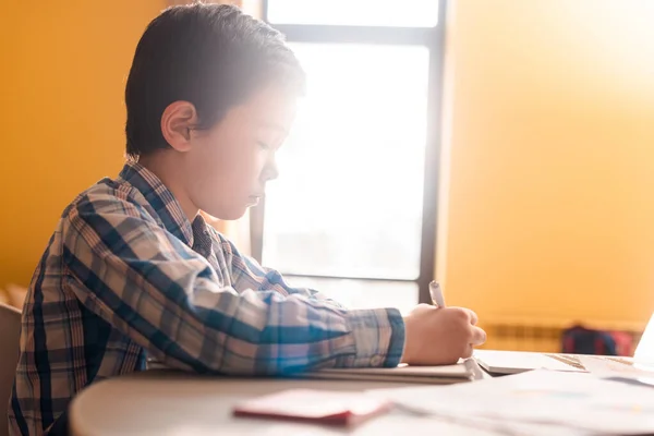 Мальчик пишет и учится дома при солнечном свете во время карантина — стоковое фото