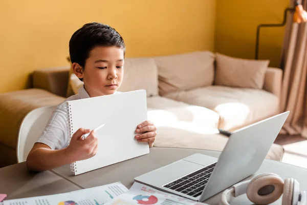 Chico asiático estudiando en línea con notepad y portátil en casa durante el autoaislamiento. - foto de stock