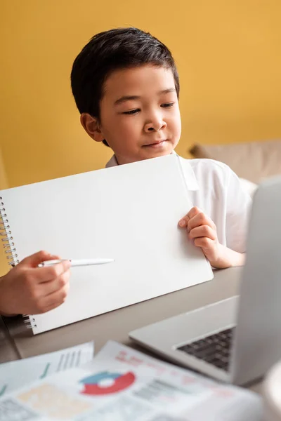 Chico asiático lindo estudiando en línea con notepad y portátil en casa durante la cuarentena. - foto de stock