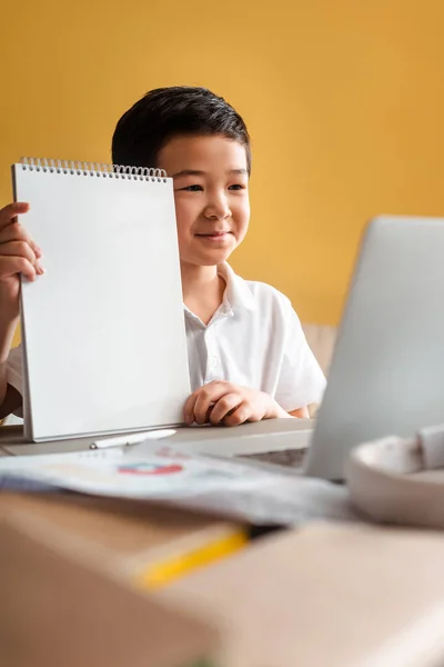 Garçon asiatique heureux étudiant en ligne avec bloc-notes et ordinateur portable à la maison pendant la quarantaine — Photo de stock
