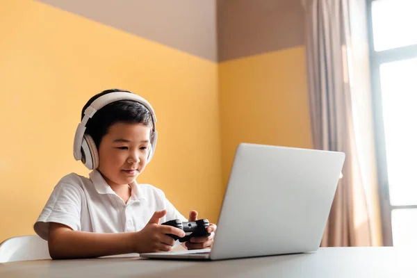 KIEV, UKRAINE - 22 AVRIL 2020 : un garçon asiatique joue à un jeu vidéo avec une manette de jeu et un ordinateur portable en auto-isolement — Photo de stock