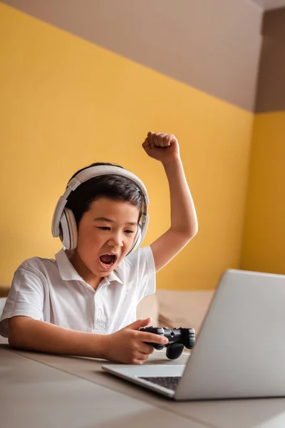 KYIV, UKRAINE - APRILE 22, 2020: ragazzo asiatico emozionale che gioca a videogiochi con joystick e laptop in quarantena — Foto stock