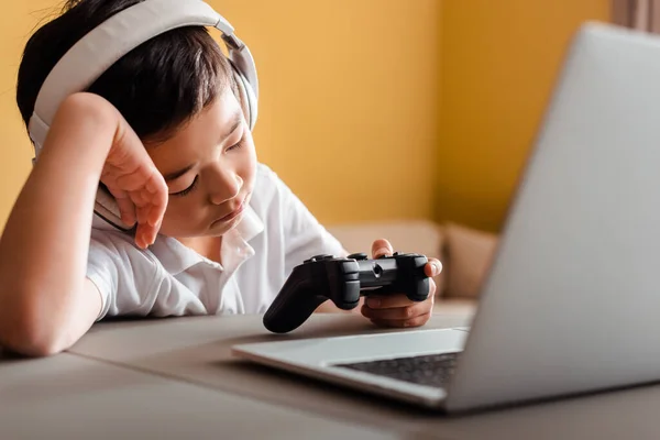 KYIV, UKRAINE - APRIL 22, 2020: sleepy asian boy playing video game with joystick and laptop on quarantine — Stock Photo