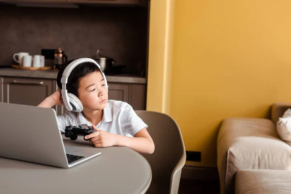 KYIV, UKRAINE - APRILE 22, 2020: noioso ragazzo asiatico che gioca a videogiochi con joystick e laptop in quarantena — Foto stock