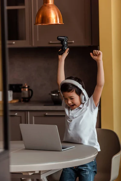 KYIV, UKRAINE - APRIL 22, 2020: jovencito asiático excitado jugando video juego con joystick y laptop en autoaislamiento. - foto de stock