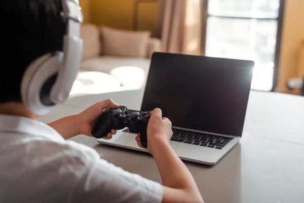 KIEV, UKRAINE - 22 AVRIL 2020 : vue en coupe d'un petit garçon jouant à un jeu vidéo avec manette de jeu et ordinateur portable avec écran vierge en auto-isolement — Photo de stock