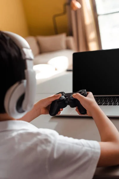 KIEV, UKRAINE - 22 AVRIL 2020 : vue en coupe d'un petit garçon jouant à un jeu vidéo avec manette de jeu et ordinateur portable avec écran vierge en quarantaine — Photo de stock